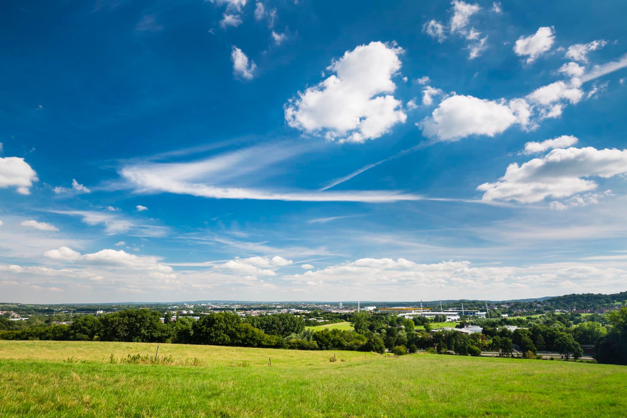 Landschaftsaufnahme einer Stadt in der Distanz, im Vordergrund sieht man grüne Wiese und einen blauen Himmel mit Wolken