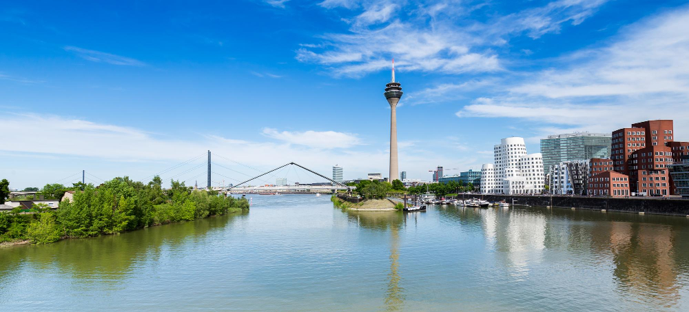 Stadtansicht von Düsseldorf mit einer Brücke, einem Turm und Gebäuden, im Vordergrund fließt der Rhein, der Himmel ist blau