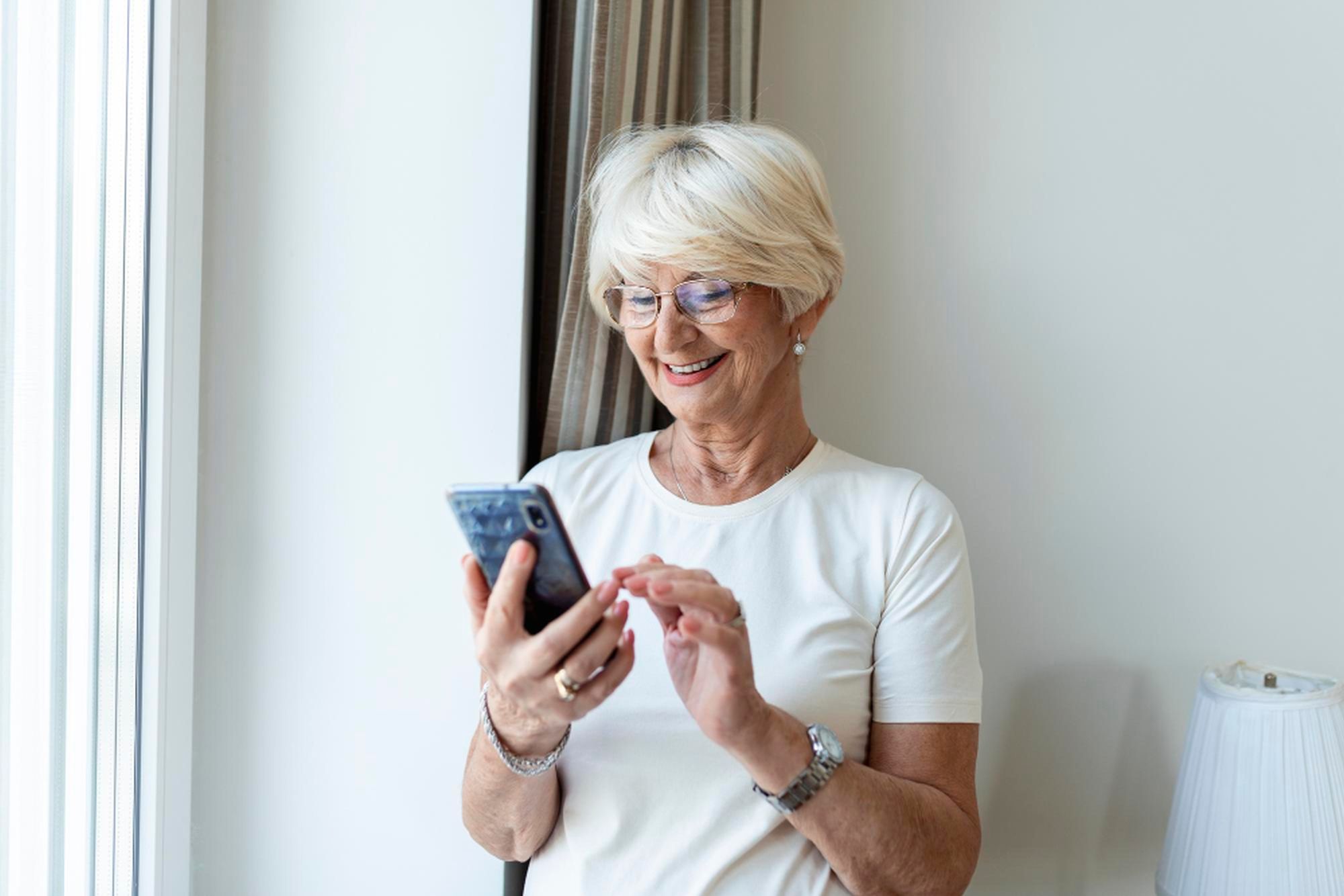 Eine ältere Dame steht vor einer weißen Wand in ihrer Wohnung und blickt auf ihr Smartphone