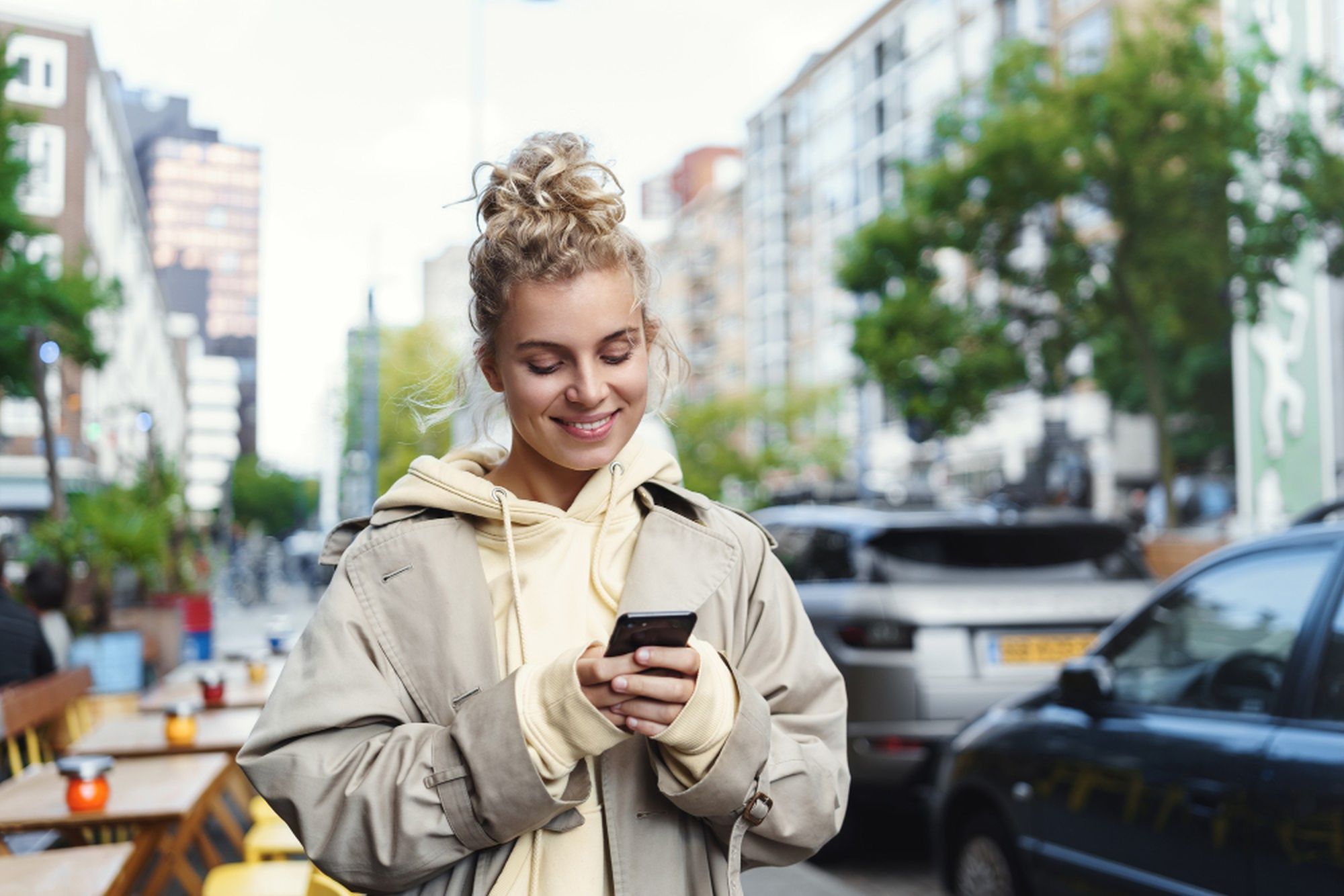 Eine junge Frau schaut auf ihr Handy, im Hintergrund sind Häuser, Autos und Bäume verschwommen zu sehen