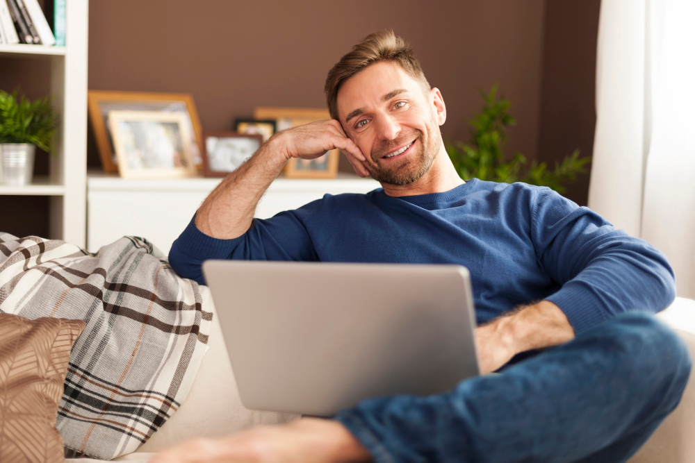 Ein Mann sitzt auf einem Sofa mit dem aufgeklappten Laptop auf dem Schoß und lächelt in die Kamera, im Hintergrund sieht man Wohnungseinrichtung wie Regale und Bilder.