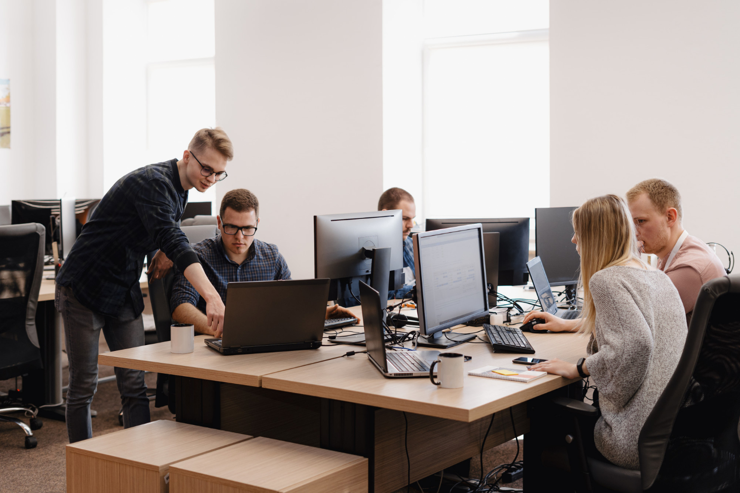 An zwei Bürotischen sitzen vier Männer und eine Frau und arbeiten an ihren Computern, ein Mann steht und zeigt auf den Bildschirm eines Computers