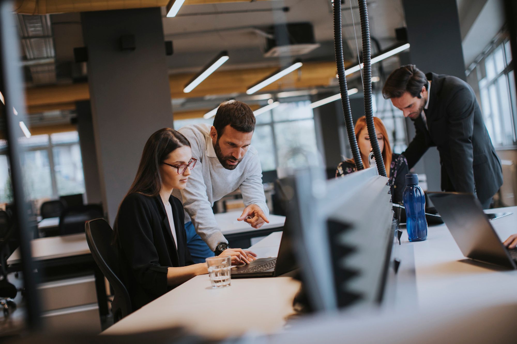 Eine Frau sitzt an einem Bürotisch, ein Mann beugt sich zu ihr und zeigt auf ihren Laptop, im Hintergrund sieht man zwei weitere Mitarbeitende und verschwommen das Büro