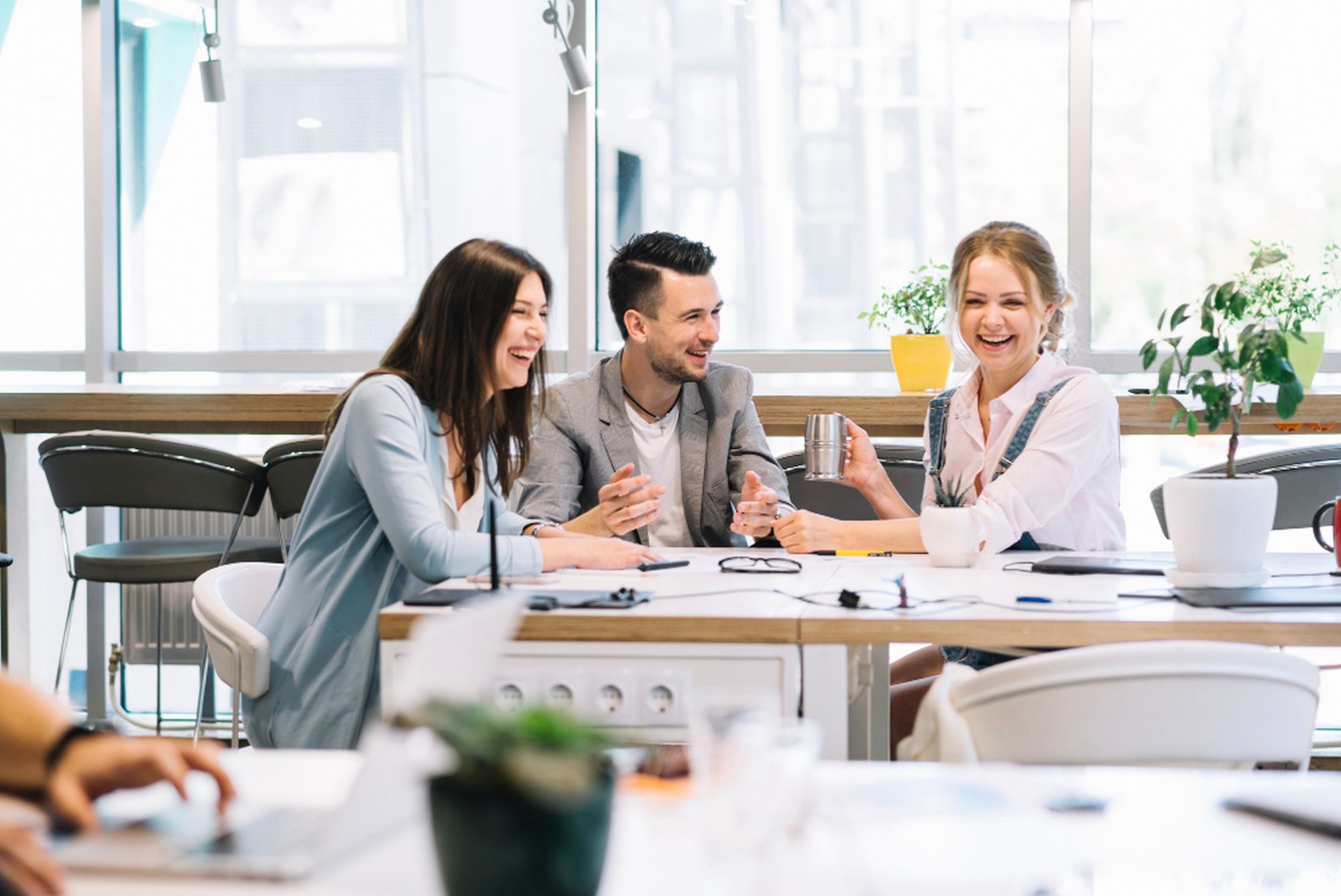 Zwei Frauen und ein Mann sitzen gemeinsam an einem Tisch in einem Büro und lachen, im Hintergrund sind Fenster, ein Tisch und Stühle zu sehen