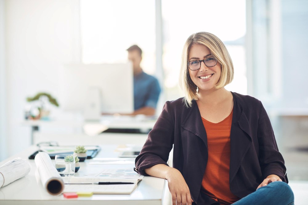 Frau, die an einem Schreibtisch sitzend freundlich in die Kamera lächelt, im verschwommenen Hintergrund arbeitet ein Mann an seinem Computer