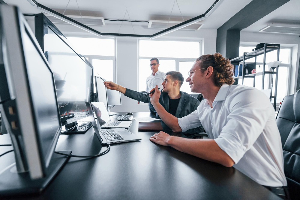 Zwei Männer sitzen in einem Büro an dunklen Schreibtischen, zeigen auf Monitore und reden, im Hintergrund steht ein Mann und schaut ihnen zu.