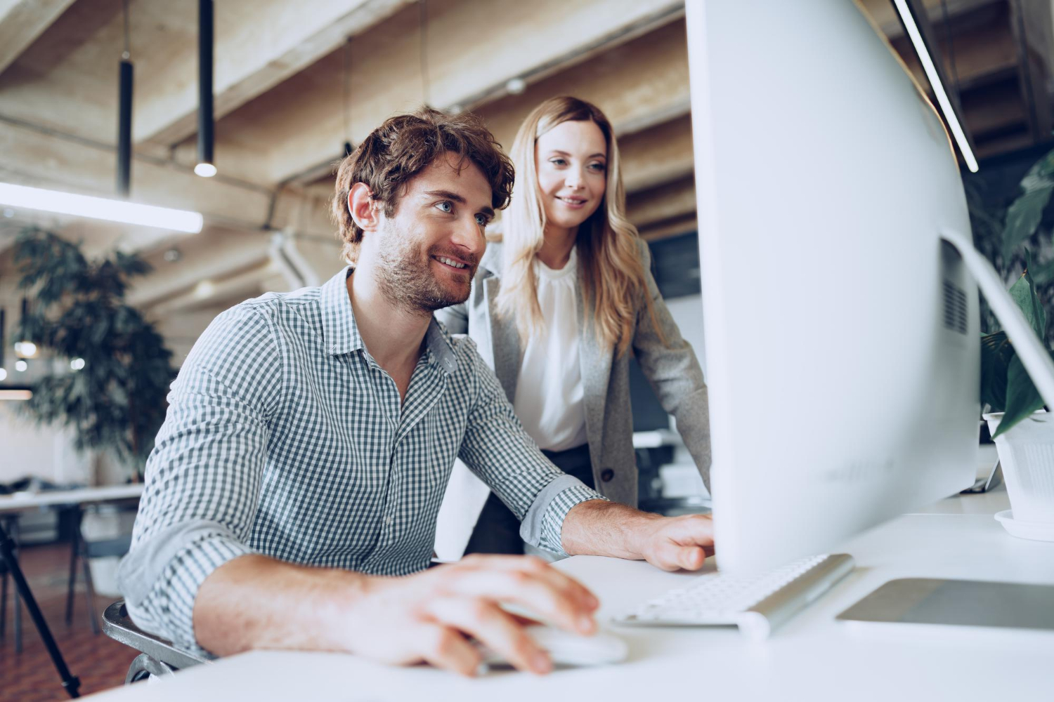 Ein Mann und eine Frau beugen sich in einem Büro über den Bildschirm eines Computers, der Mann sitzt am Schreibtisch, die Frau steht neben ihm