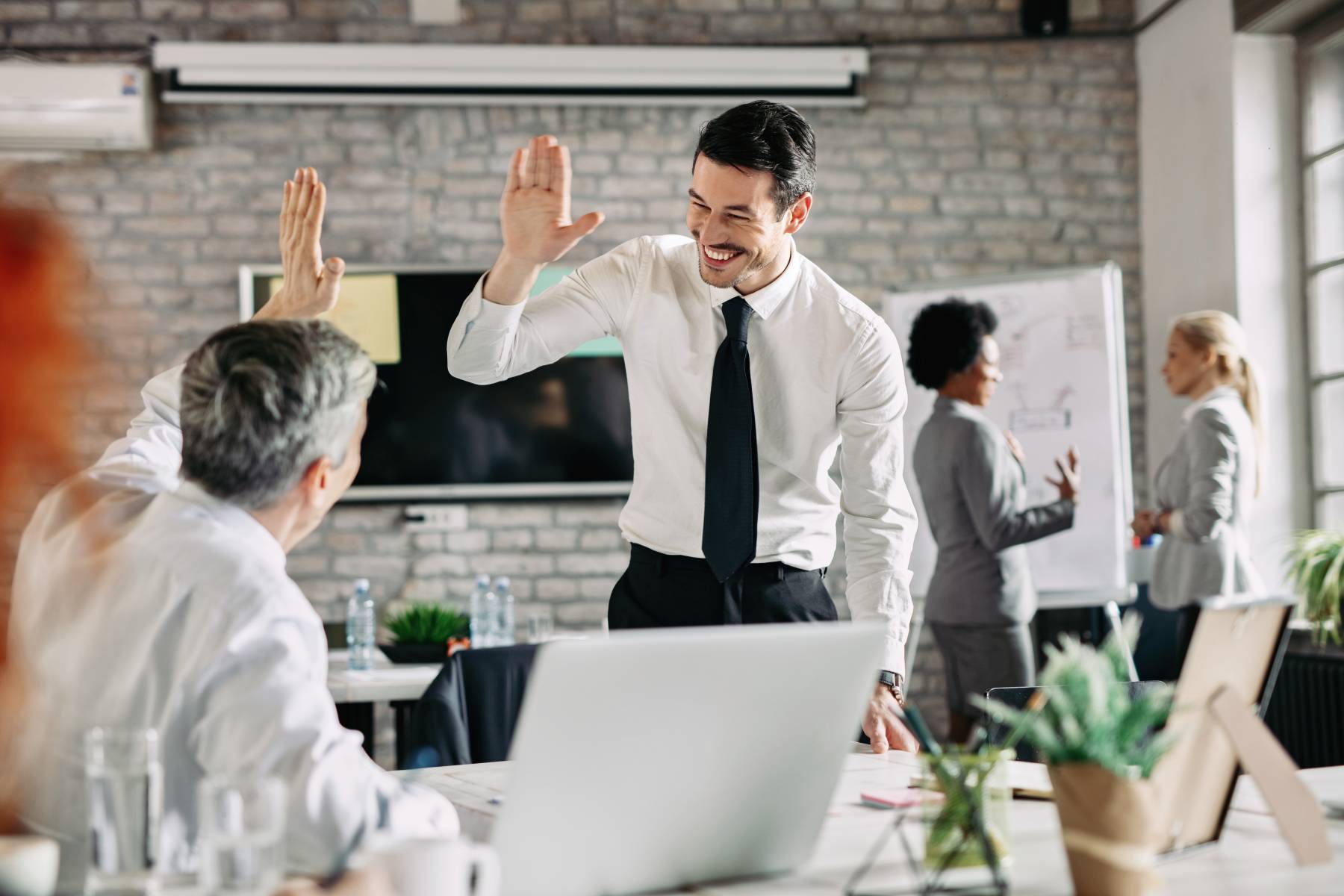Zwei Männer in Anzügen geben sich in einem Büro High Five, im Hintergrund diskutieren zwei Damen mit einander