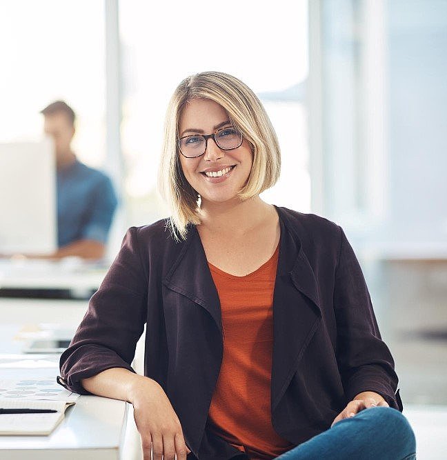 Frau, die an einem Schreibtisch sitzend freundlich in die Kamera lächelt, im verschwommenen Hintergrund arbeitet ein Mann an seinem Computer