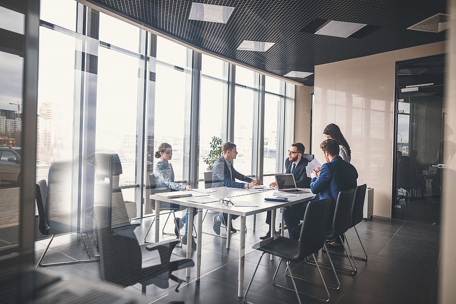 Blick durch ein großes Fenster in einen Konferenzraum, mehrere Mitarbeitende sitzen an einem Tisch und diskutieren, hinter ihnen ist eine große Fensterfront