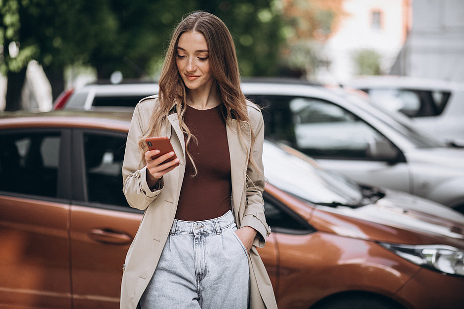Eine junge Frau steht vor einen geparkten Auto und schaut auf ihr Handy, im Hintergrund sind etwas verschwommen weitere Autos und ein Baum zu sehen.