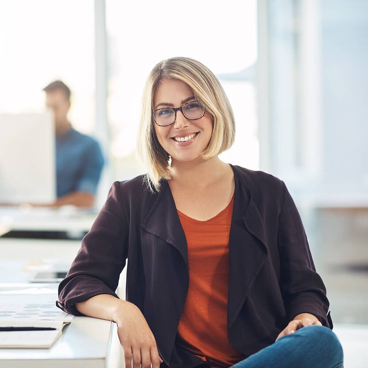 Frau, die an einem Schreibtisch sitzend freundlich in die Kamera lächelt, im verschwommenen Hintergrund arbeitet ein Mann an seinem Computer