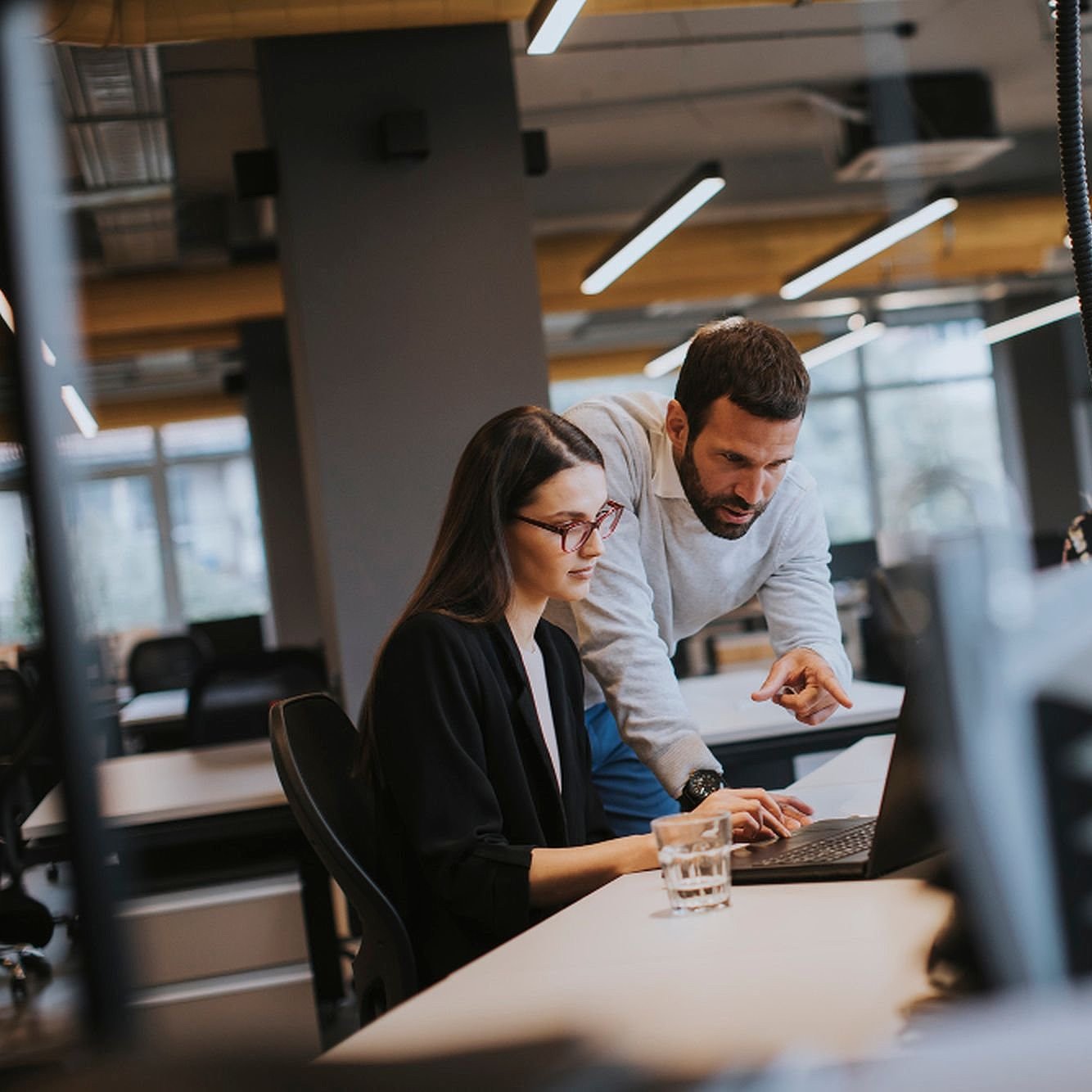 Eine Frau sitzt an einem Bürotisch, ein Mann beugt sich zu ihr und zeigt auf ihren Laptop, im Hintergrund sieht man zwei weitere Mitarbeitende und verschwommen das Büro