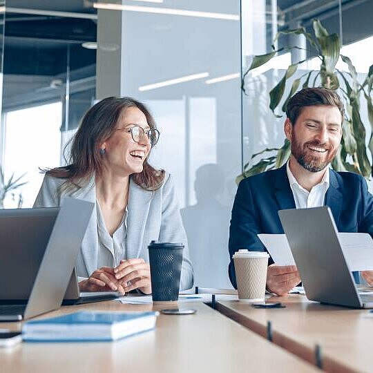 Zwei Männer und zwei Frauen sitzen an eine Konferenztisch, reden und Lachen, zwei Laptops, Unterlagen und Kaffeebecher stehen auf dem Tisch