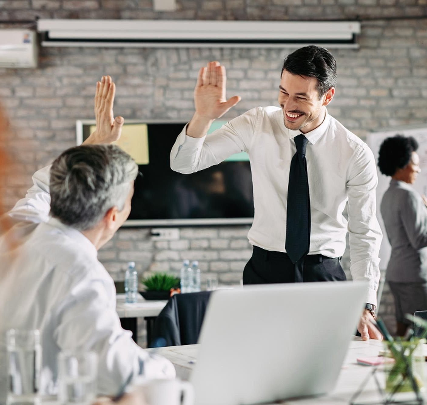 Zwei Männer in Anzügen geben sich in einem Büro High Five, im Hintergrund diskutieren zwei Damen mit einander