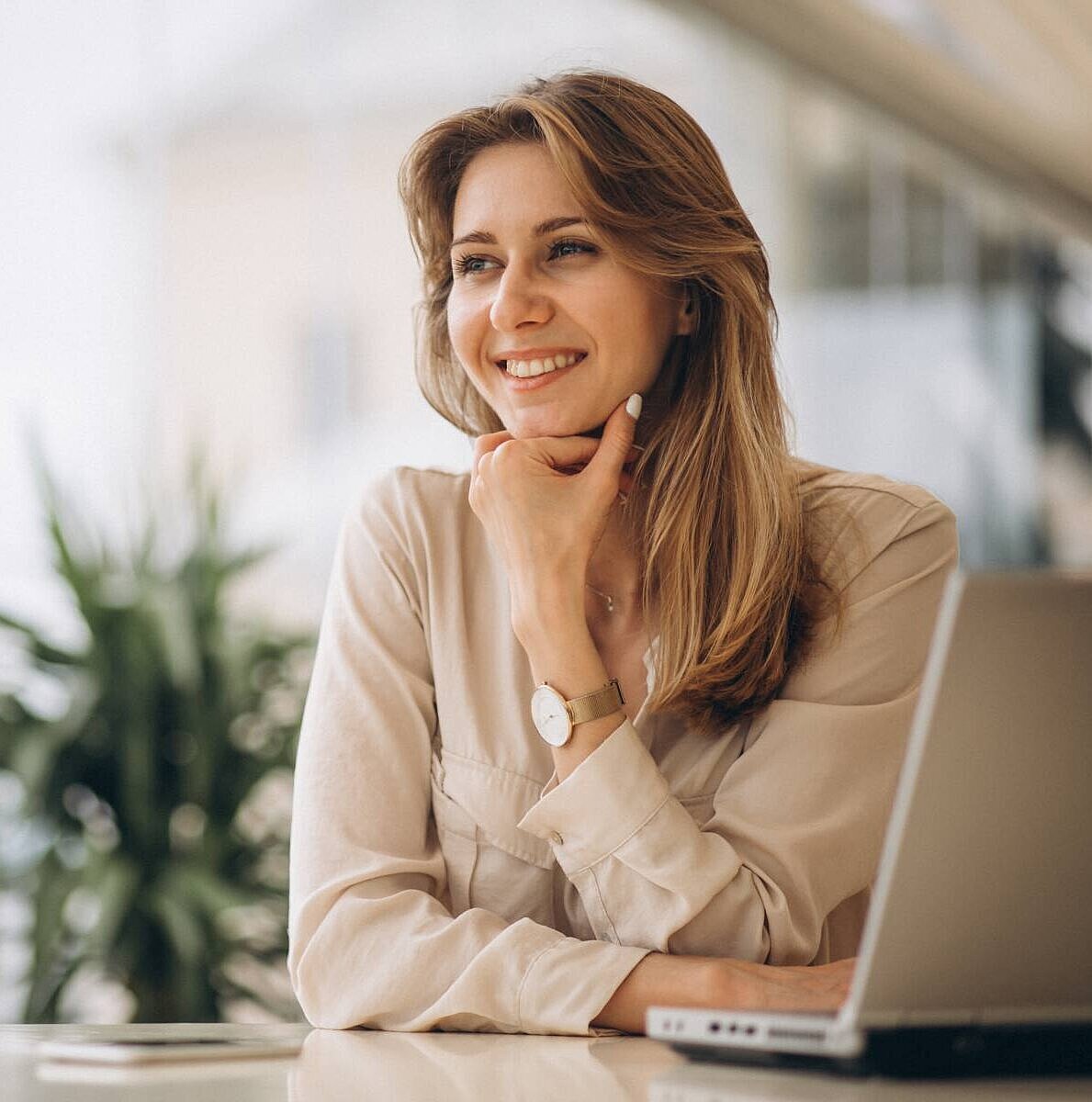 Eine Frau sitzt an einem Tisch, stützt sich auf ihren Unterarmen ab und lächelt, neben ihr steht ein aufgeklappter Laptop