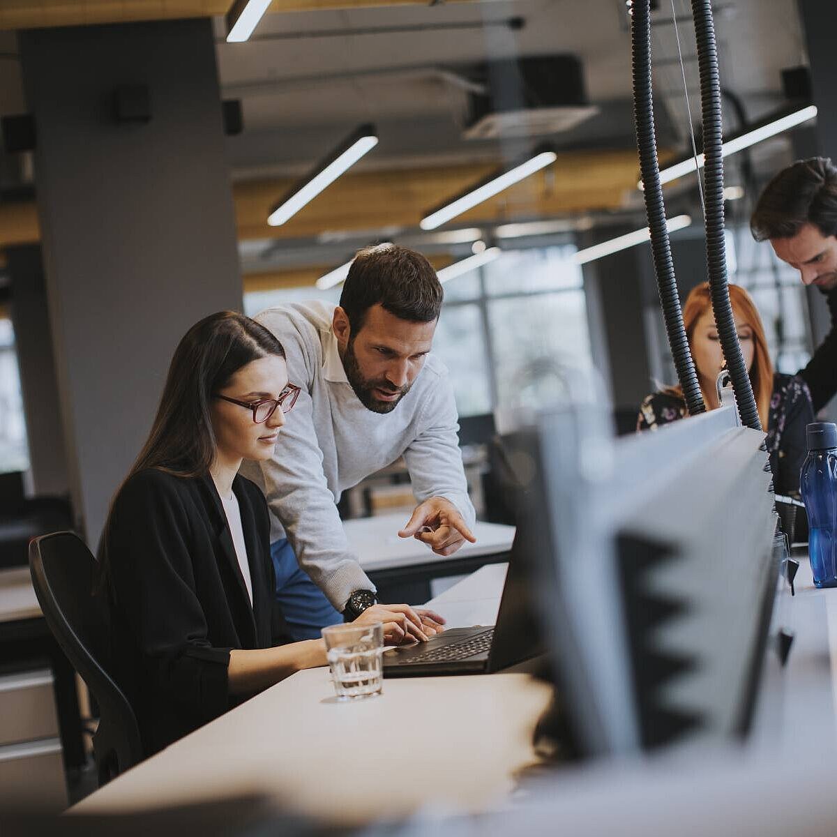 Eine Frau sitzt an einem Bürotisch, ein Mann beugt sich zu ihr und zeigt auf ihren Laptop, im Hintergrund sieht man zwei weitere Mitarbeitende und verschwommen das Büro