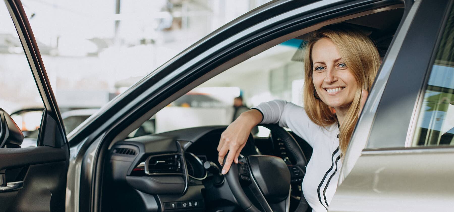 Eine Frau sitzt seitlich auf dem Fahrersitz eines Autos und lächelt in die Kamera, die Autotür ist geöffnet