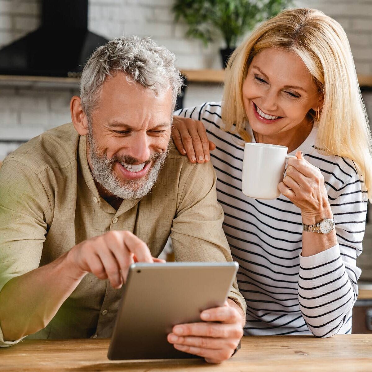 Ein mittelaltes Pärchen steht in seiner Küche und beugt sich lächelnd über ein Tablet, die Dame hält eine Tasse in der Hand