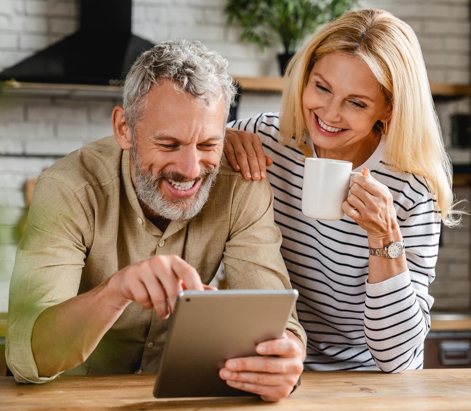 Ein mittelaltes Pärchen steht in seiner Küche und beugt sich lächelnd über ein Tablet, die Dame hält eine Tasse in der Hand