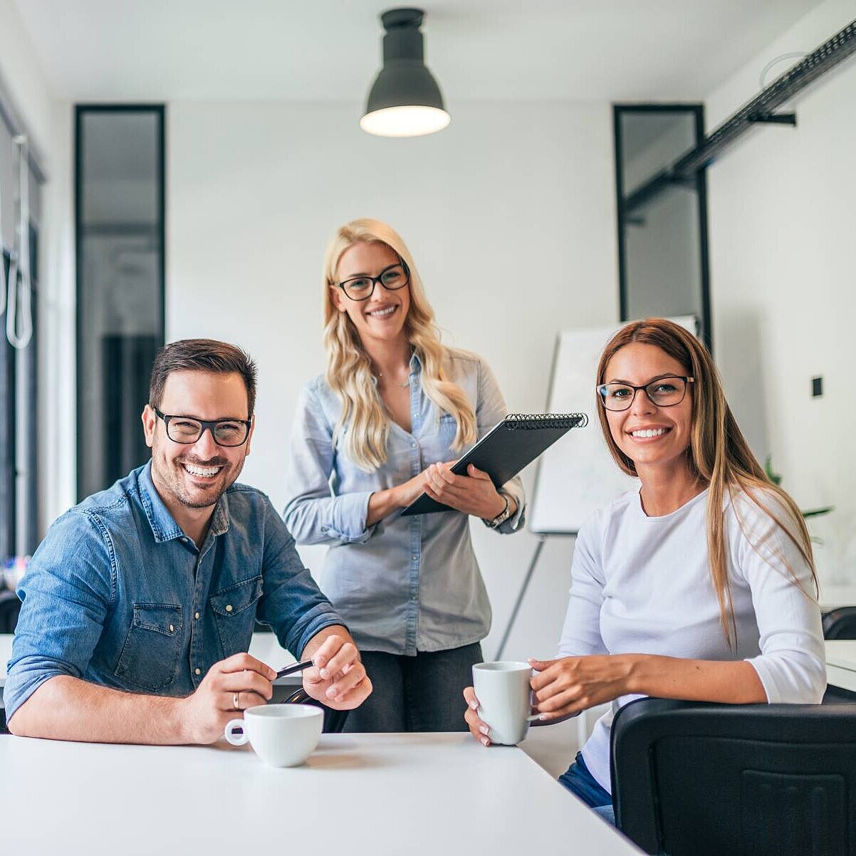 Ein junger Mann und eine junge Frau sitzen an einem Schreibtisch, hinter ihnen steht eine weitere Frau, sie lächeln in die Kamera
