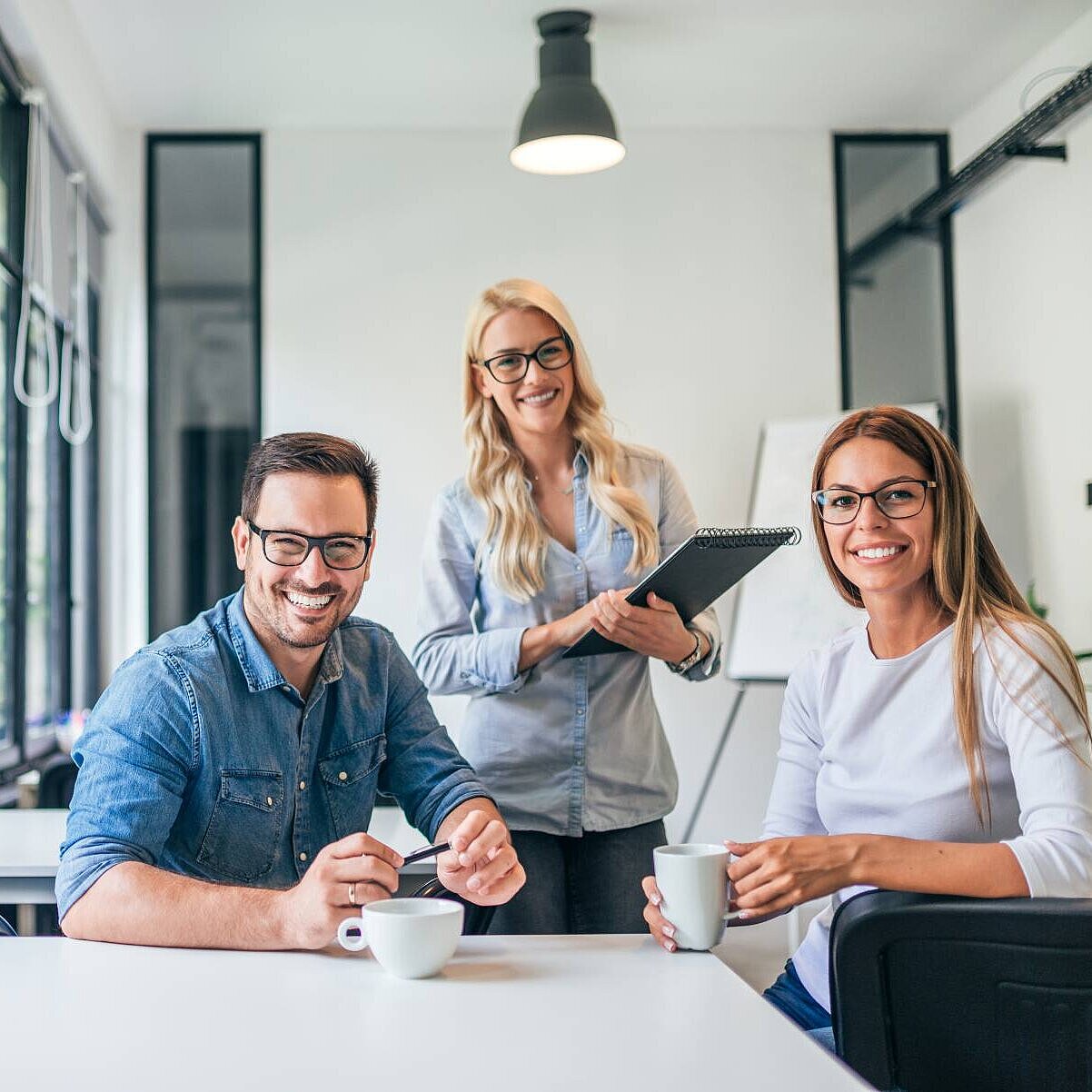 Ein junger Mann und eine junge Frau sitzen an einem Schreibtisch, hinter ihnen steht eine weitere Frau, sie lächeln in die Kamera