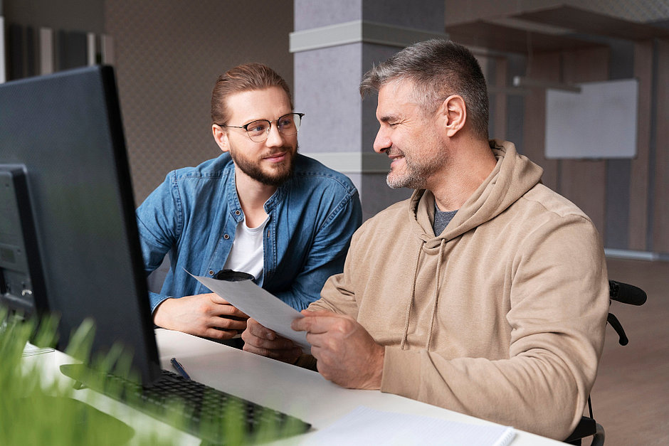 Zwei sich zugewandte Männer an einem Schreibtisch mit Computer im Dialog, einer hält ein Blatt Papier in den Händen