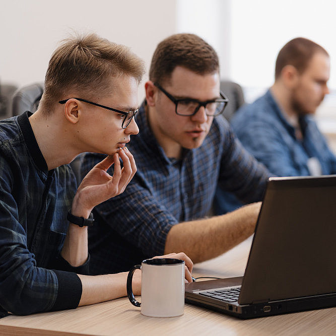 Zwei Männer sitzen an einem Bürotisch und schauen sich grübelnd etwa auf einem Laptop an, im Hintergrund sitzt ein weiterer Mann vor seinem Computer