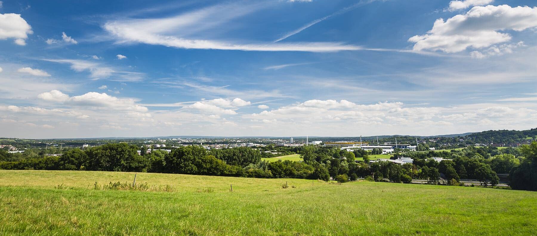 Landschaftsaufnahme einer Stadt in der Distanz, im Vordergrund sieht man grüne Wiese und einen blauen Himmel mit Wolken