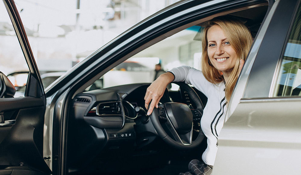 Eine Frau sitzt seitlich auf dem Fahrersitz eines Autos und lächelt in die Kamera, die Autotür ist geöffnet