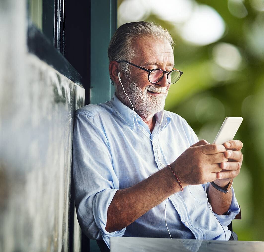 Ein älterer Herr lehnt an der Wand eines Gebäudes und blickt lächelnd auf sein Smartphone, im Hintergrund sind verschwommen Bäume zu sehen