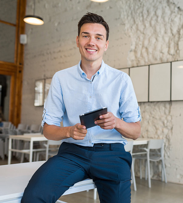 Junger Mann der lächelnd auf der Kante eines Schreibtisches sitzt und ein Tablet in der Hand hält